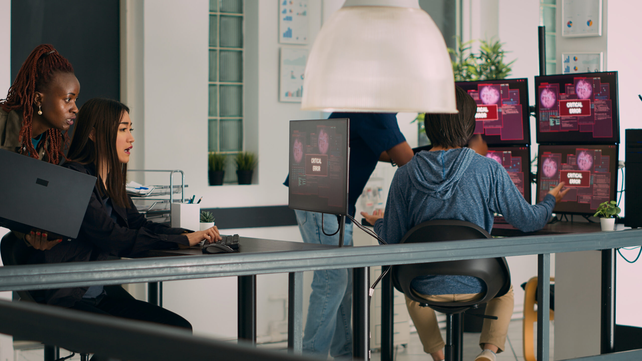 Multiethnic team of coders encountering system error message on computer, trying to solve hacking alert problem. Diverse developers having hacked server on multiple monitors. Tripod shot.